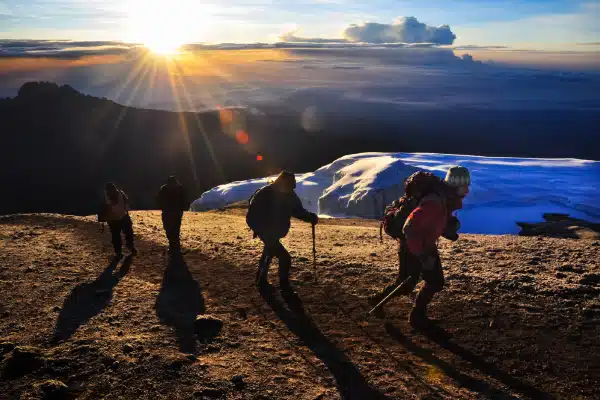 Four hikers with backpacks and walking sticks trek uphill on a rugged, sunlit mountain slope with a snow-covered area in the background, as the sun rises, casting long shadows.