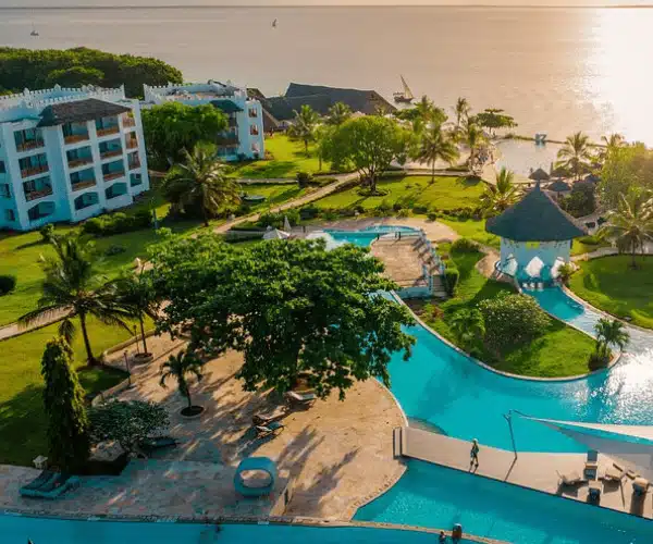 Aerial view of a tropical beach resort with a large swimming pool, palm trees, white buildings, green lawns, and the ocean in the background under a golden sunset.